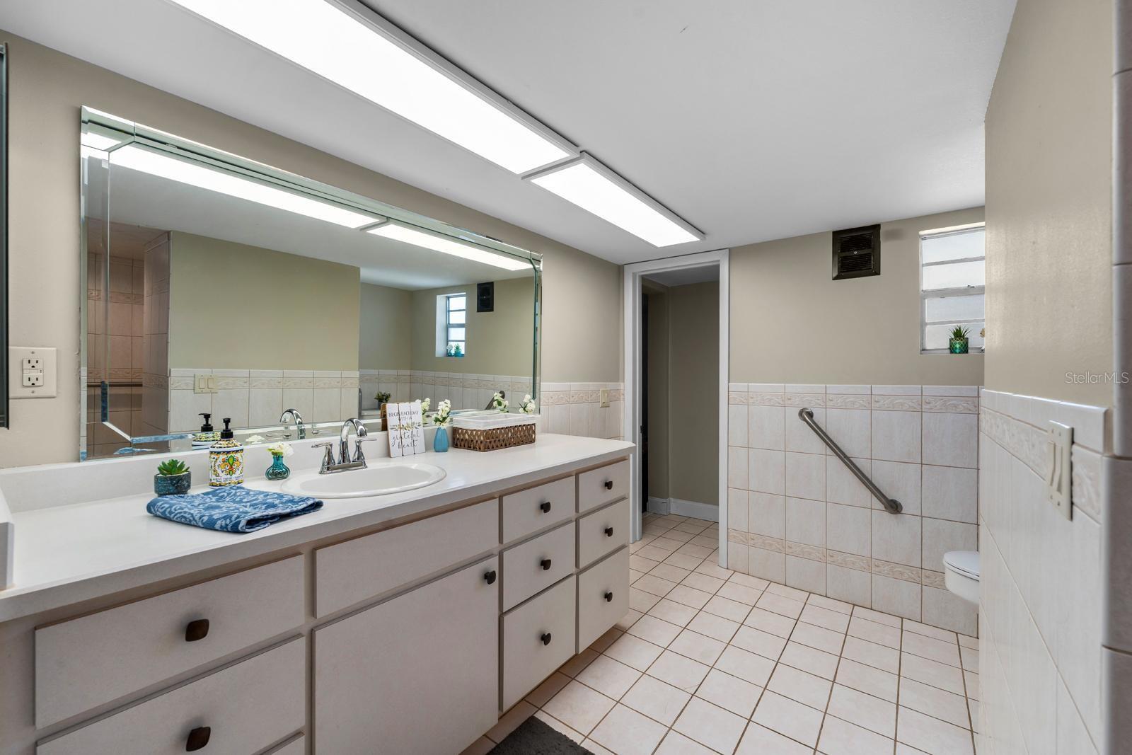 The bathroom features a tiled walk-in shower with grab bars and a built-in bench, ceramic tile flooring, a mirrored wall cabinet, a walk-in closet, newer countertop, tile on the walls (and two grab bars by the commode), fresh paint, and new lighting.