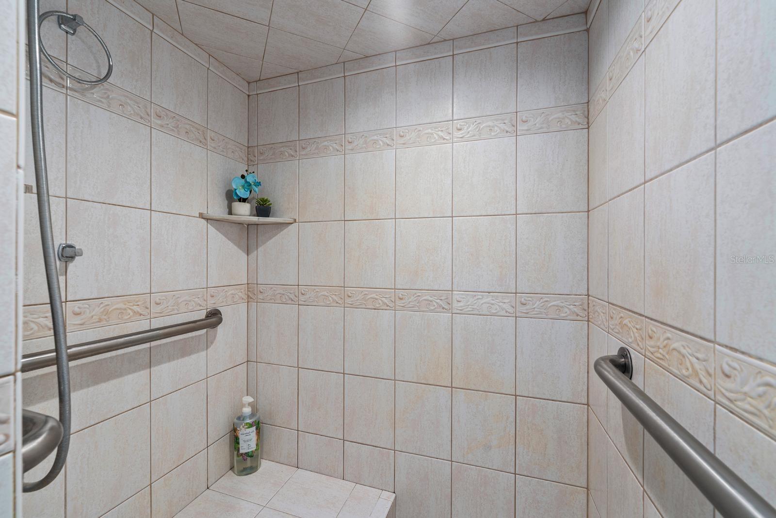 The bathroom features a tiled walk-in shower with grab bars and a built-in bench, ceramic tile flooring, a mirrored wall cabinet, a walk-in closet, newer countertop, tile on the walls (and two grab bars by the commode), fresh paint, and new lighting.