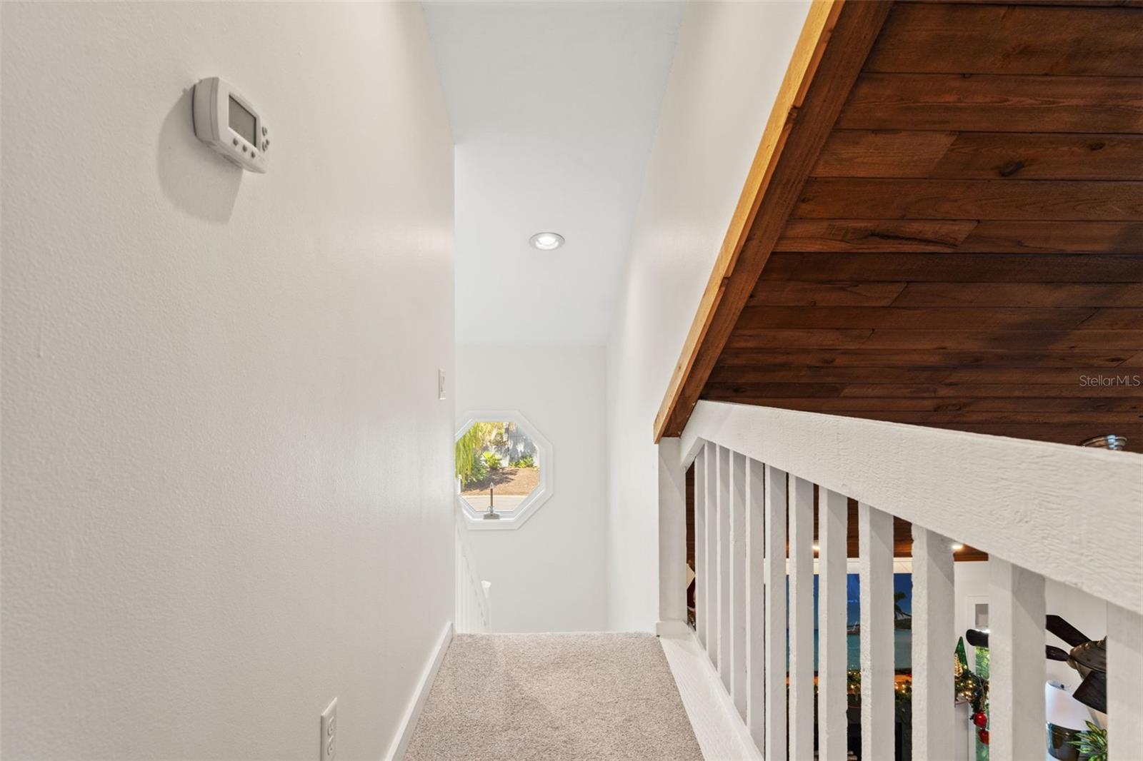 Upper-level hallway with carpeted flooring, white railing, and natural light from an octagonal window.