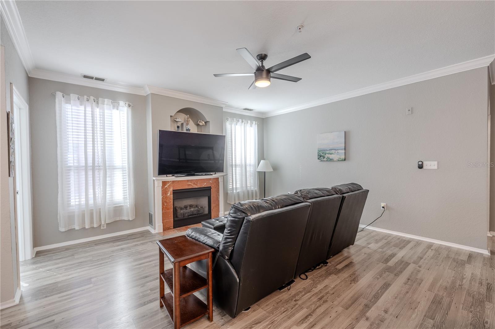 This inviting living area is anchored by a gas fireplace with a classic mantle and decorative niche above, flanked by tall windows with sheer curtains that fill the space with soft, natural light.