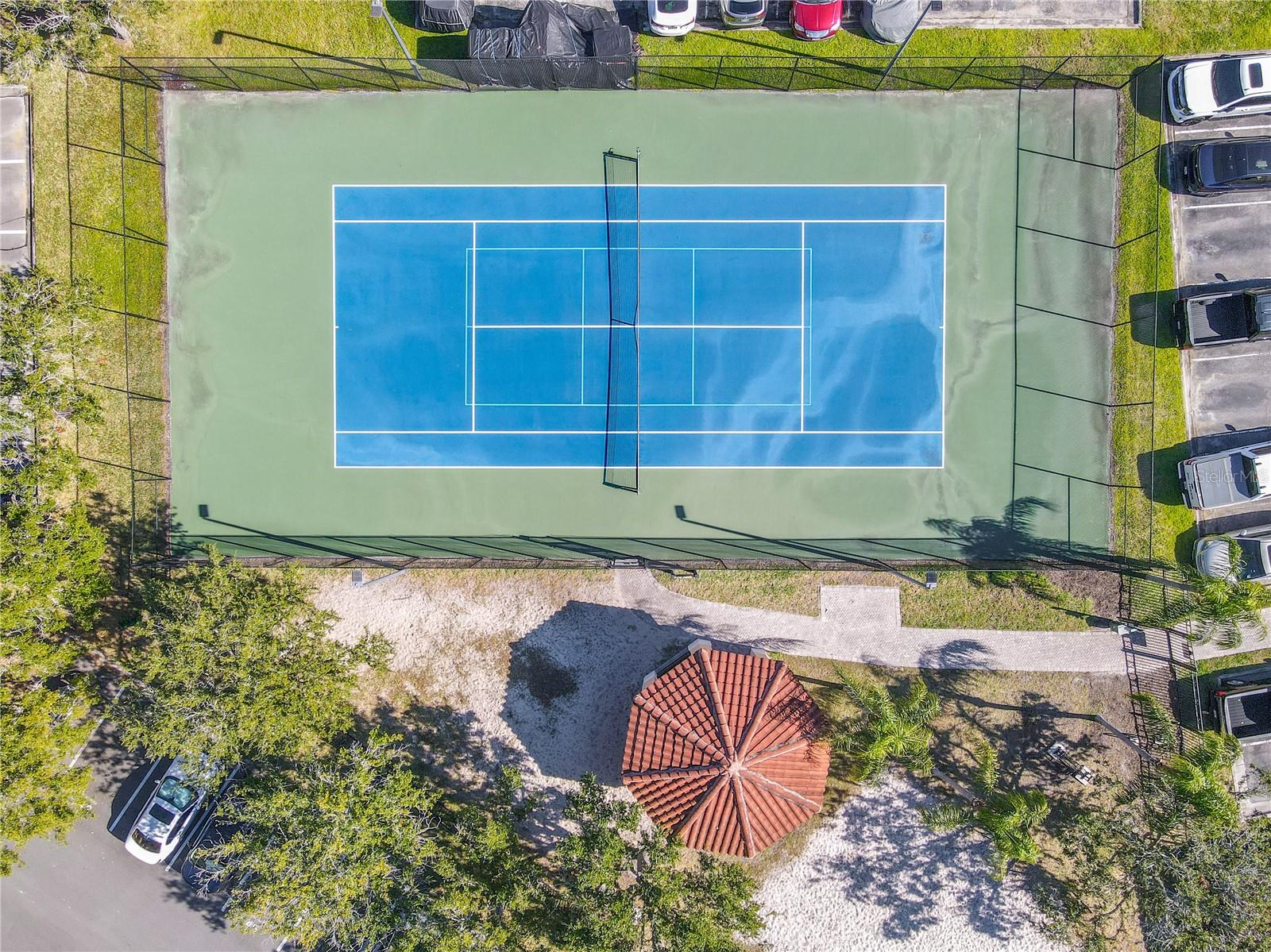 A tranquil community courtyard with a shaded pavilion, sandy play area, and palm-lined green space, framed by elegant Mediterranean-style residences under clear blue skies.