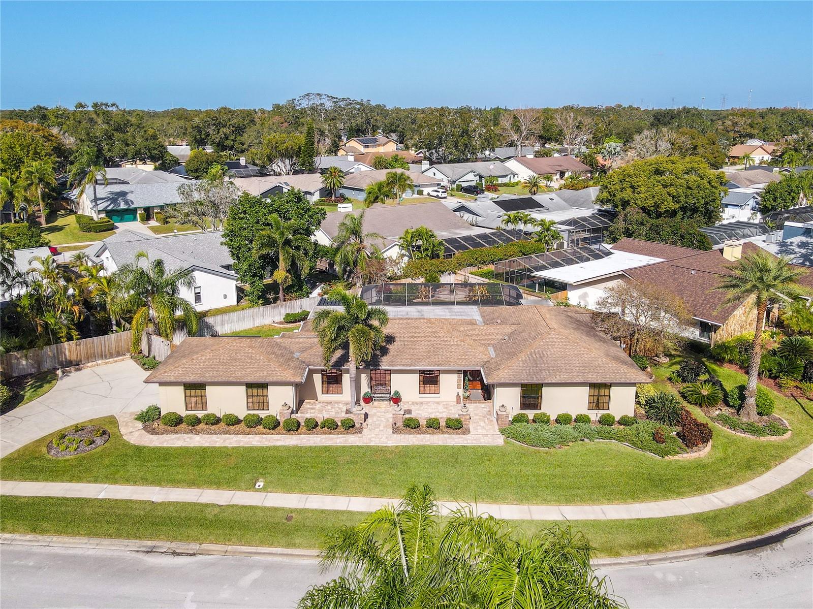 A beautifully maintained single-story home set on a wide, manicured lot, featuring a long curved driveway, lush tropical landscaping, a symmetrical façade, fully guttered room, and a spacious backyard oasis framed by mature palms and established neighborhood greenery.