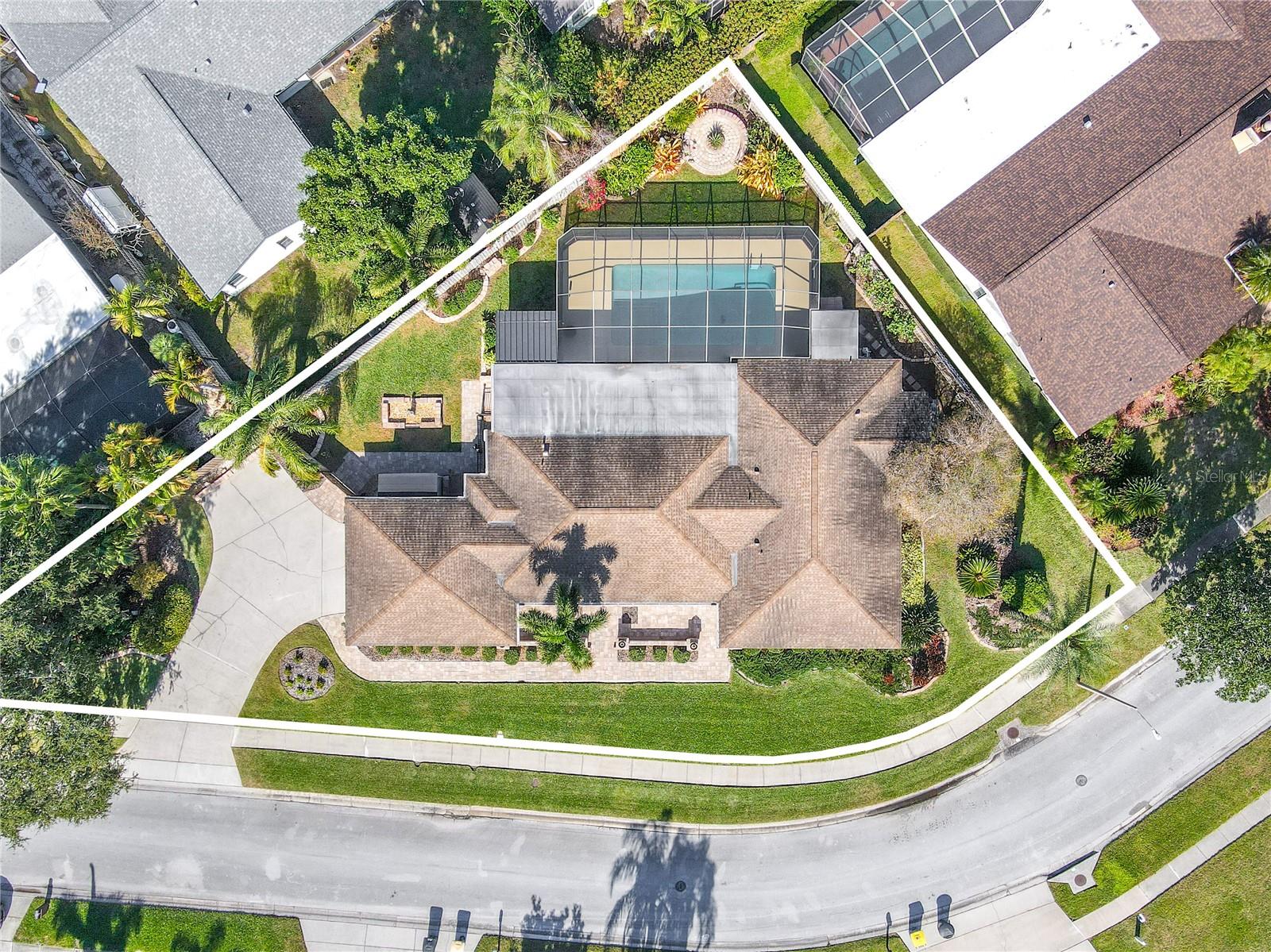 A top-down aerial view showcasing the home’s expansive, fully fenced lot with a screened pool enclosure, thoughtfully designed paver patios and walkways, a large driveway with ample parking, and lush tropical landscaping that maximizes privacy and outdoor living space.