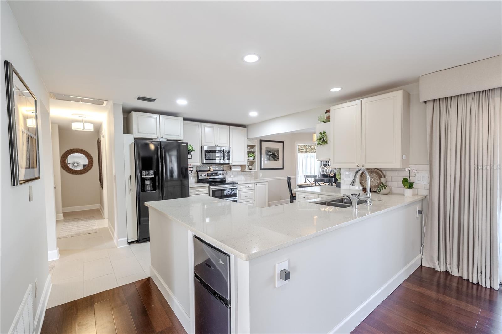 View of the bright, open kitchen from the family room, highlighting expansive quartz countertops, a large center island with seating, crisp white cabinetry, recessed lighting, and seamless flow into the main living space.