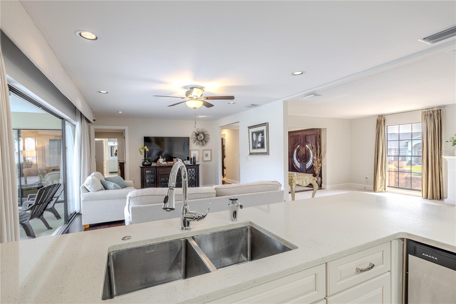 An open-concept view from the quartz kitchen peninsula highlights seamless flow into the family room, formal living room and foyer, featuring recessed lighting, a ceiling fan, expansive sight lines, and abundant natural light ideal for entertaining.