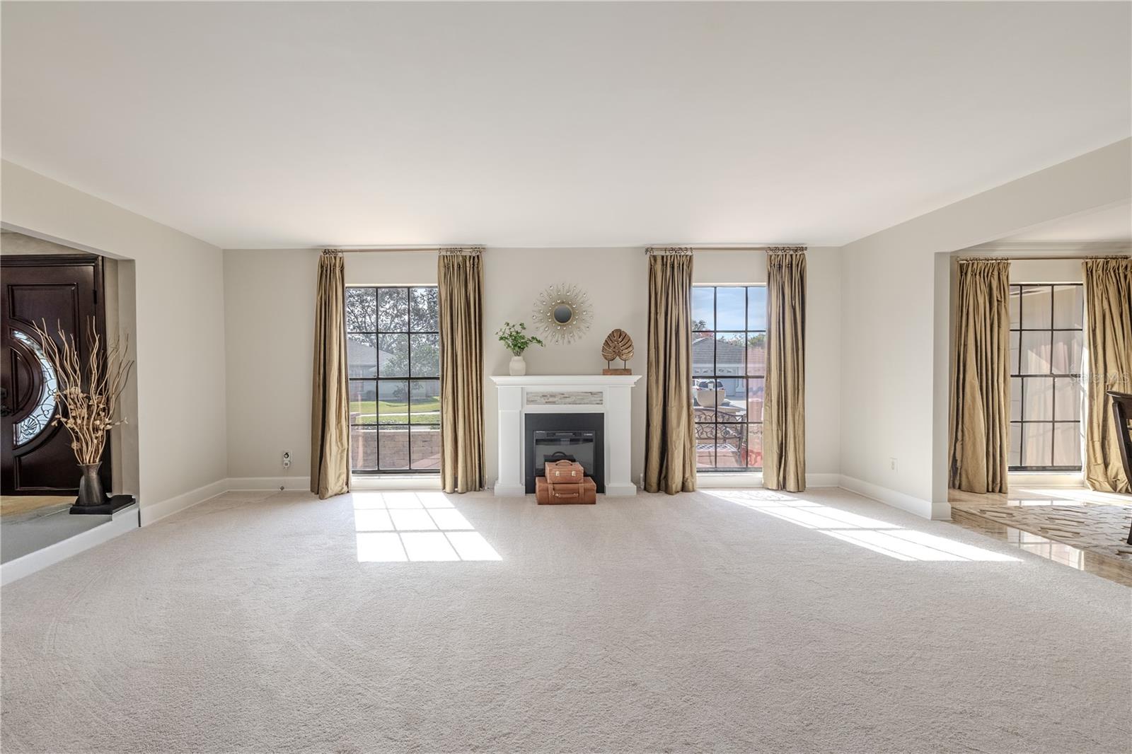 A bright formal living room anchored by a classic fireplace features floor-to-ceiling windows that flood the space with natural light and provide seamless access to the adjoining formal dining room.