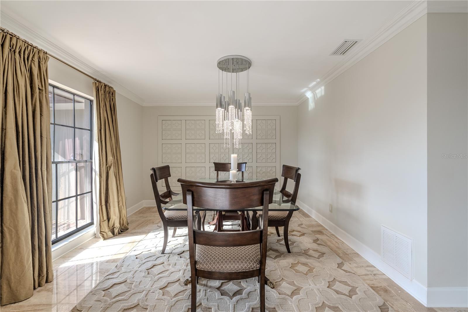 The formal dining room features elegant porcelain tile flooring, custom wall detailing, crown moulding, and a statement chandelier, with natural light streaming through floor length window dressed in floor-length drapery.