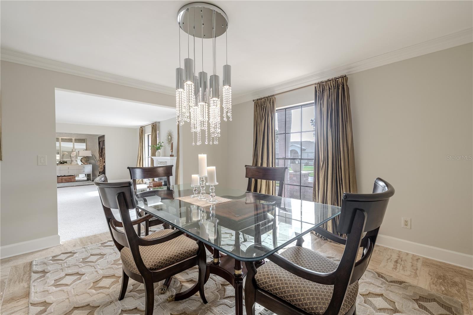 The formal dining room showcases porcelain tile flooring, crown moulding, floor-length custom drapery, and a striking crystal chandelier, creating a bright, elegant space that flows seamlessly into the formal living area.