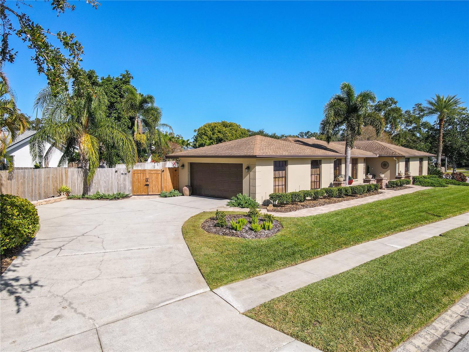 A wide-angle exterior view highlighting the home’s generous curved driveway, side-entry garage, double gate entry to the back yard, and neatly trimmed tropical landscaping that frames the property with excellent curb appeal and a clean, welcoming streetscape.