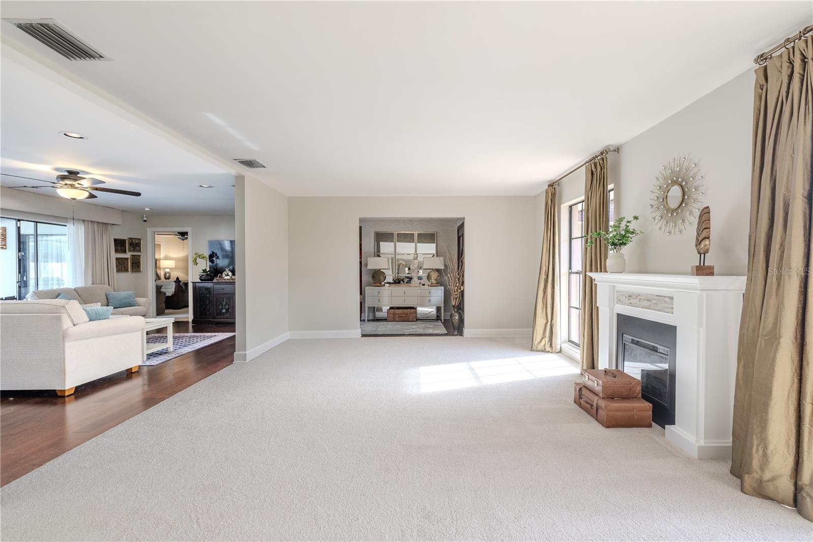View from the formal dining room into the spacious living area highlights the open layout, plush carpeting, elegant fireplace, and abundant natural light flowing through floor-length windows.