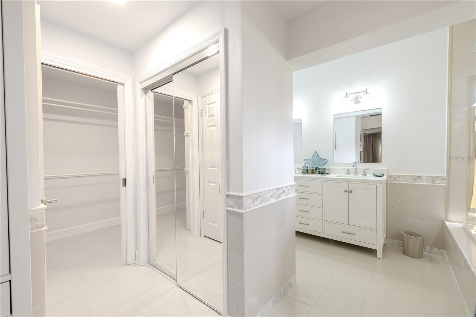 The bright primary bathroom features a clean, spa-like design with a spacious walk-in closet, additional built in closet, dual-sink vanity with ample storage, elegant tile wainscoting, light neutral large format tile finishes, and a well-lit layout that enhances both function and comfort.