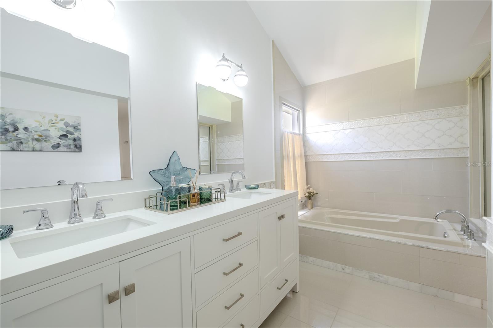 The spa-inspired primary bathroom features a double-sink vanity with sleek cabinetry, vaulted ceiling, stone countertops, abundant storage, and a soaking tub accented by elegant inlayed tile detailing framed with mother of pearl tile and soft natural light.