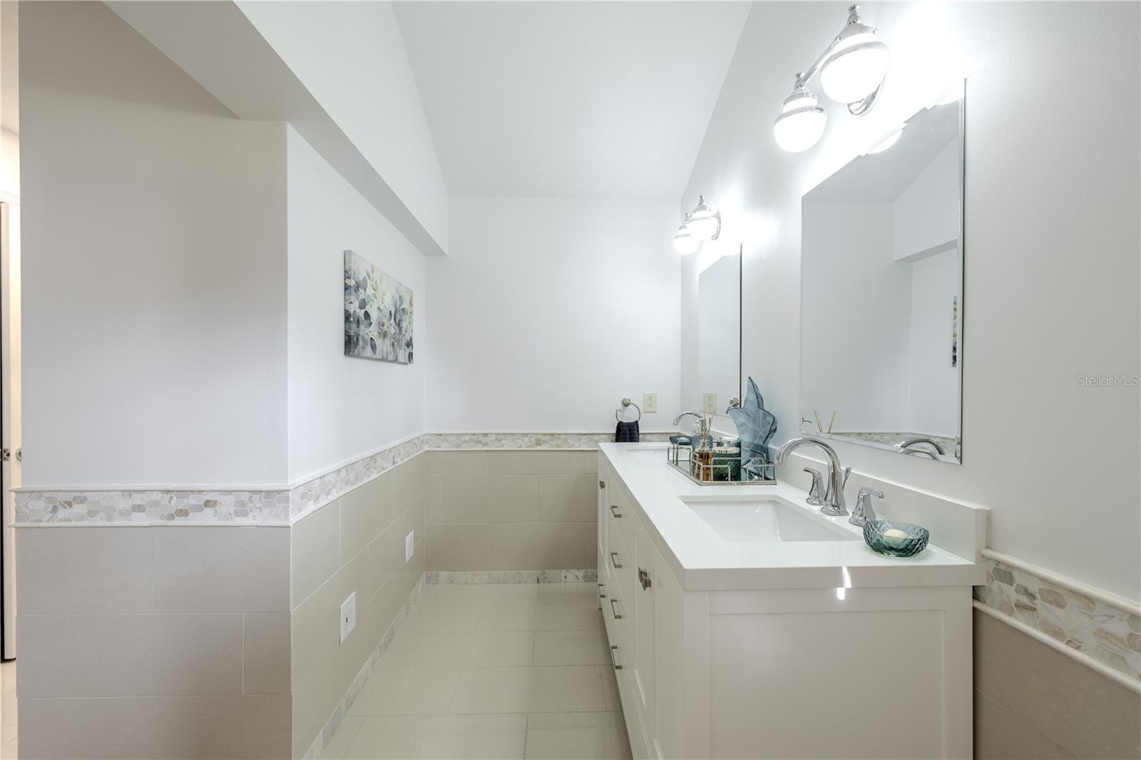 The bright, spa-like primary bathroom showcases a vaulted ceiling, long vanity with stone countertops, ample drawer storage, designer lighting, and elegant tile wainscoting that adds both texture and refinement.