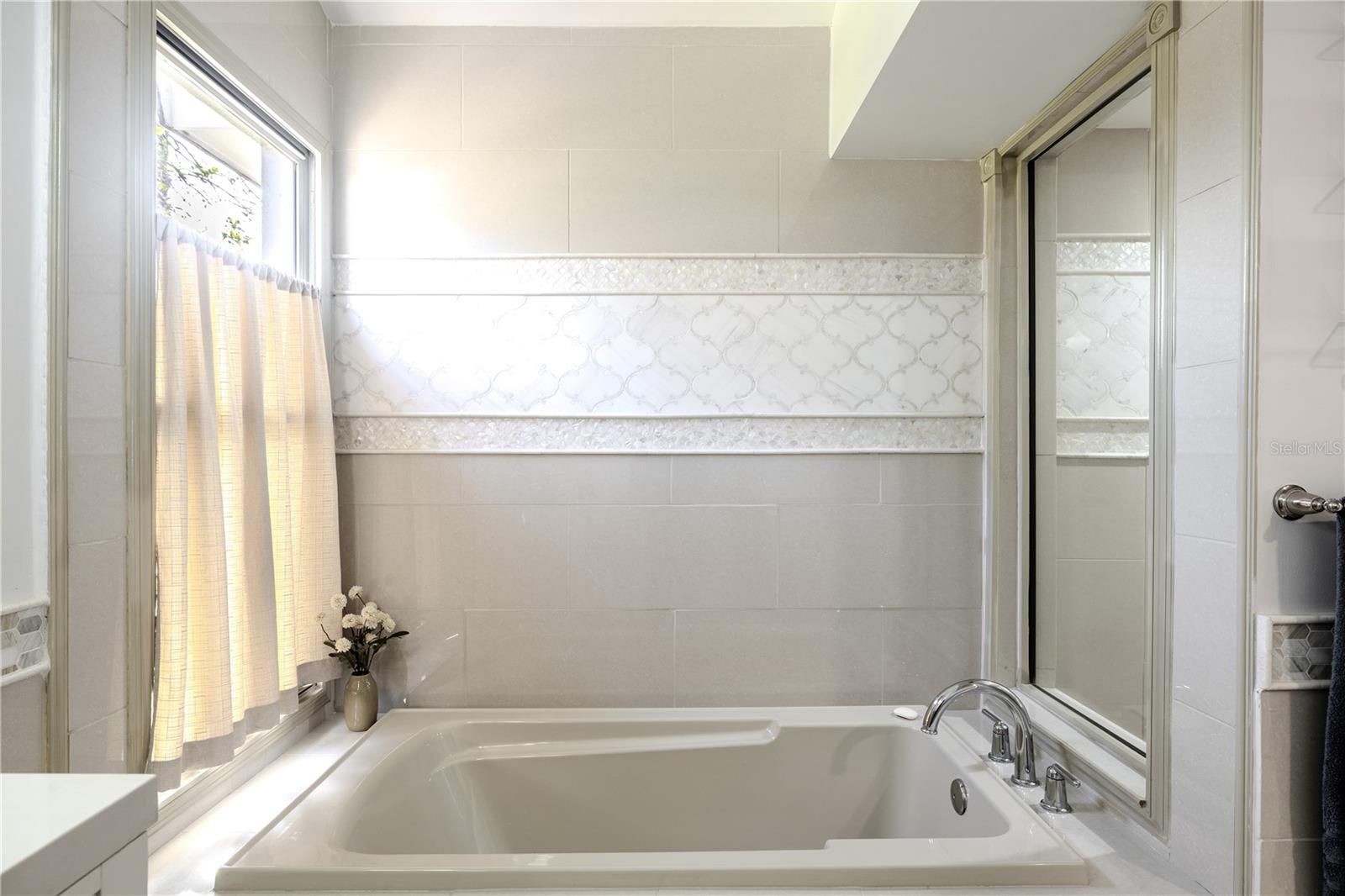 The elegant soaking tub is framed by refined tile work with decorative inlay accents, a large picture window for natural light, and a glass wall with separate shower, all with spa-inspired ambiance.
