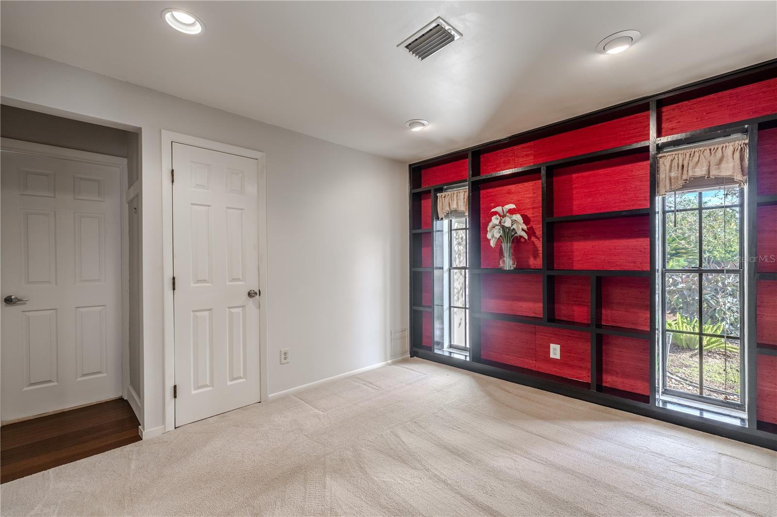 This second bedroom features plush carpeting, recessed lighting, built in closet, direct master bedroom access, and a striking custom accent wall with integrated shelving and natural light from dual windows, creating a unique and flexible space ideal for a home office, nursery, or private retreat.