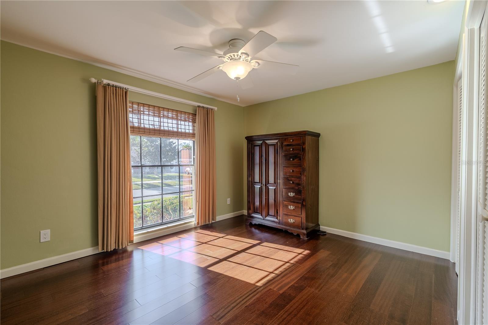 The fourth bedroom is bright and inviting, featuring rich hardwood flooring, a ceiling fan, and a large window with custom window treatments that fills the space with natural light, making it ideal as a guest room, home office, or flex space.