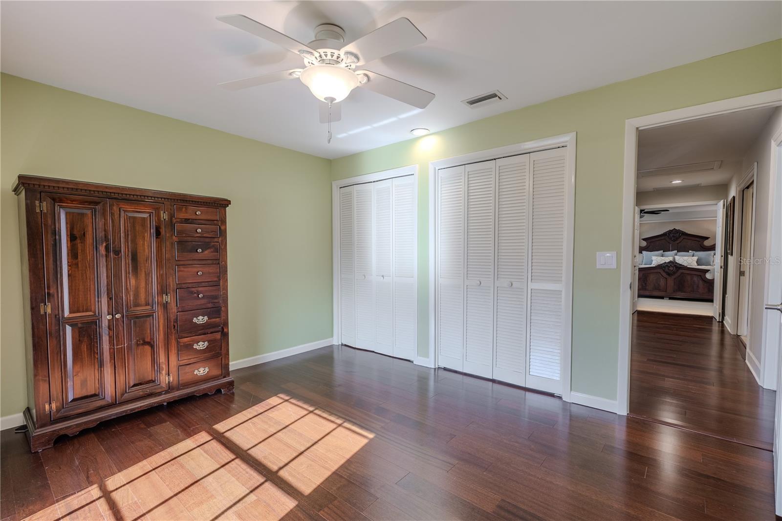 This additional view of the fourth bedroom highlights the rich hardwood flooring, ceiling fan with light, soft neutral walls, and generous dual access closets, with convenient access to the primary suite hallway—ideal for flexible living or guest accommodations.
