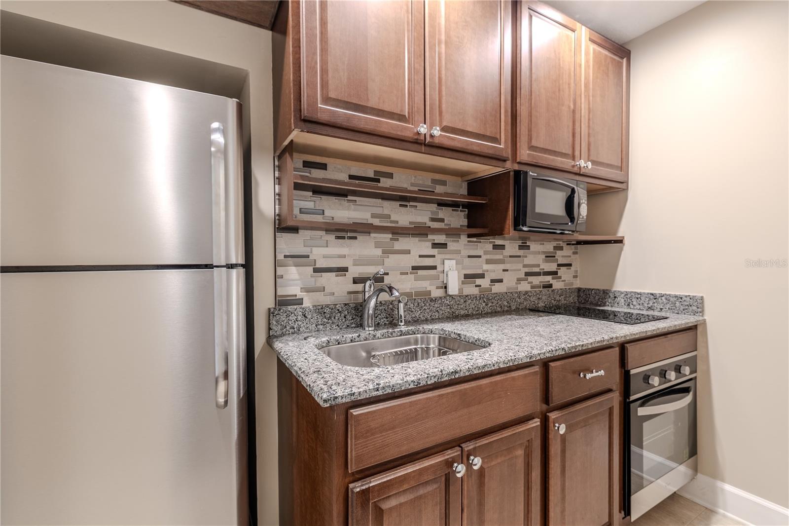 This thoughtfully designed secondary kitchen area features a convection cooktop with oven, granite countertops, custom wood cabinetry, mosaic tile backsplash, and stainless appliances—ideal for guest accommodations or multigenerational living.