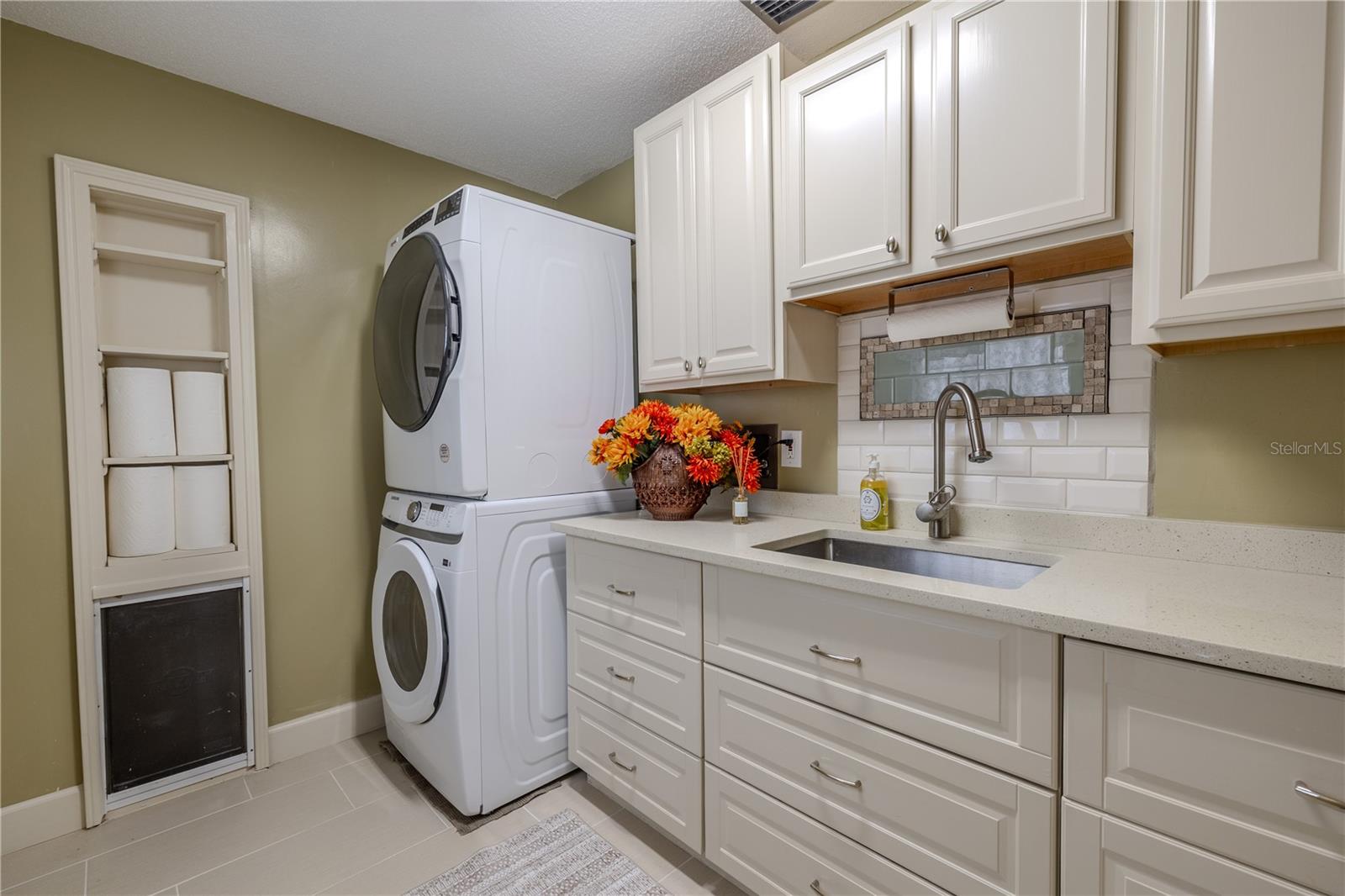 This well-appointed laundry room mirrors the main kitchen with matching cabinetry, quartz countertops, subway tile backsplash, a utility sink, and large-format tile flooring, plus a convenient oversized dog door for easy outdoor private potty area access.