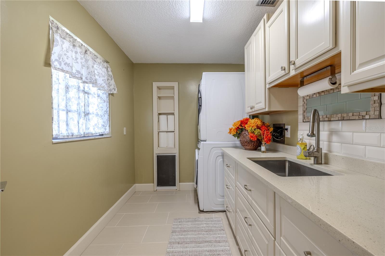 This bright and functional laundry room features matching white cabinetry, quartz countertops, a utility sink, stacked washer and dryer, large-format tile flooring, and a convenient built-in dog door, all complemented by natural light from the window.