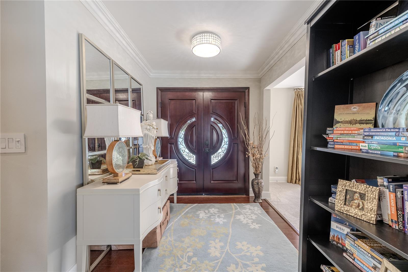 Elegant foyer entry showcasing imported solid-wood double doors with decorative glass inlays, enhanced by crown molding, refined lighting, wood floor, and a welcoming transition into the main living spaces.