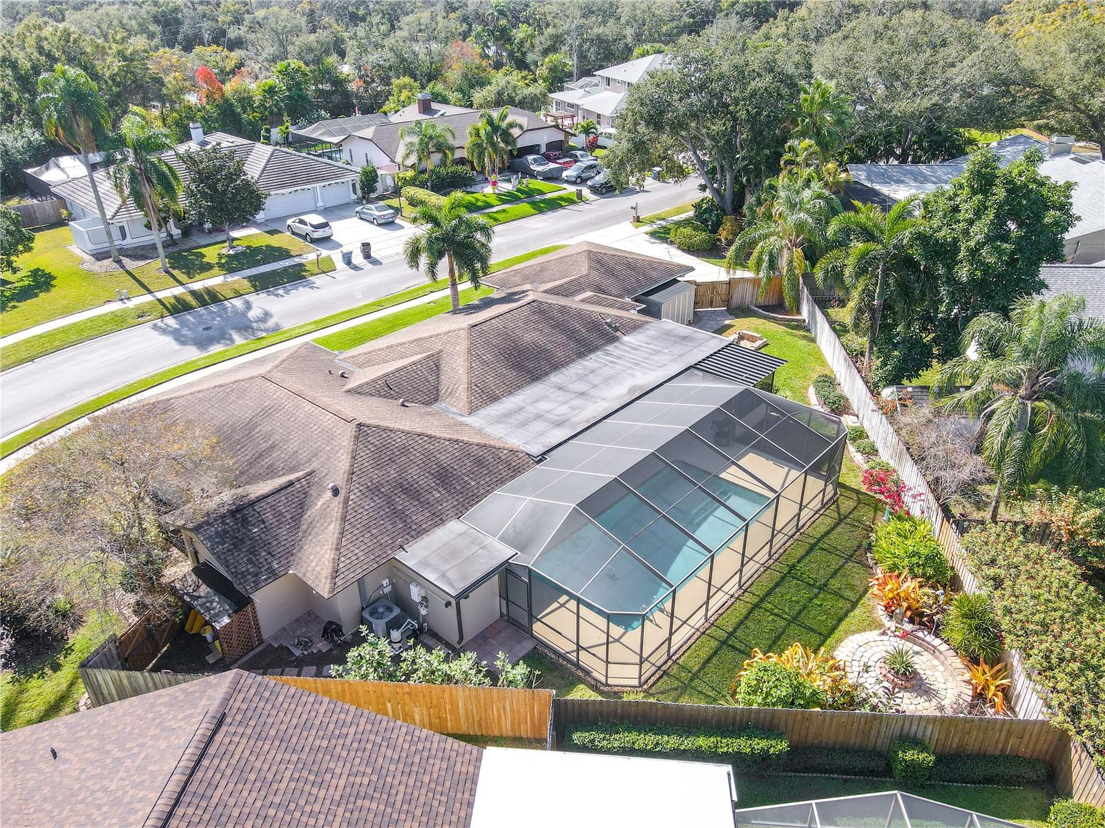 An expansive aerial backyard view highlights the home’s screened pool and lanai, private fenced backyard with mature landscaping and garden features, dual storage sheds, and a quiet residential setting with tree-lined streets and neighboring homes.