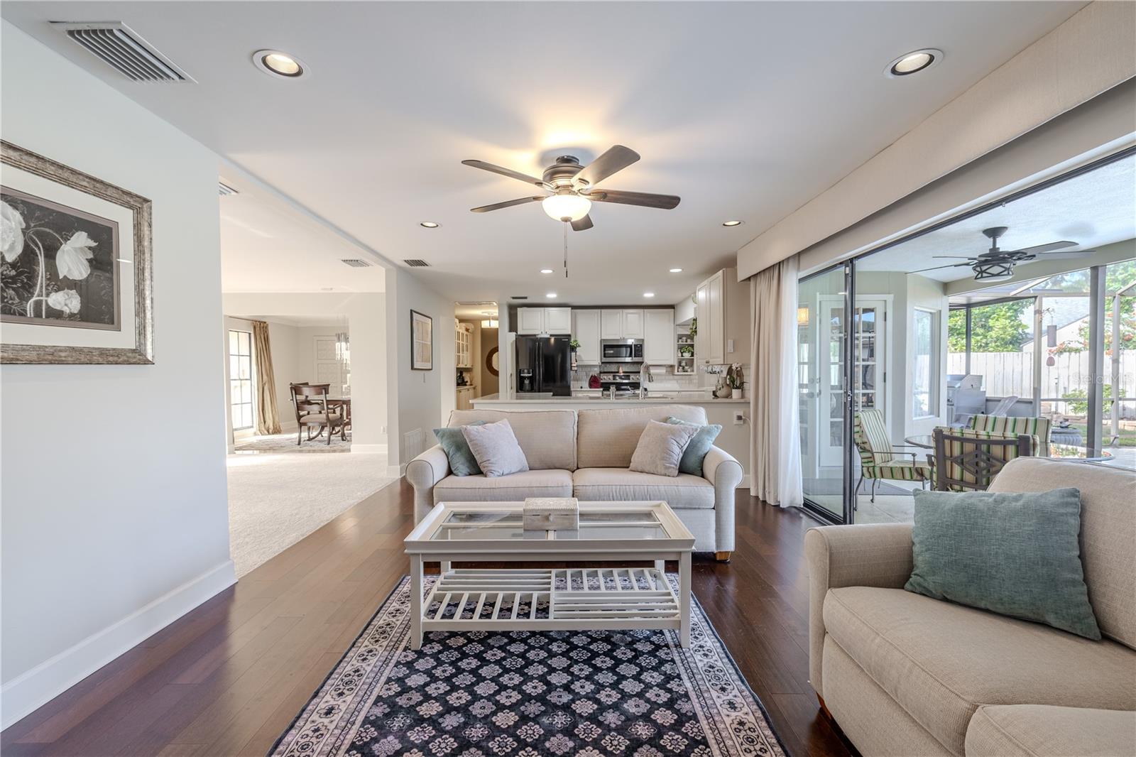 Bright, open family room featuring hardwood floors, recessed lighting, a ceiling fan, and seamless flow to the kitchen and screened outdoor lanai and pool area through expansive sliding glass doors.