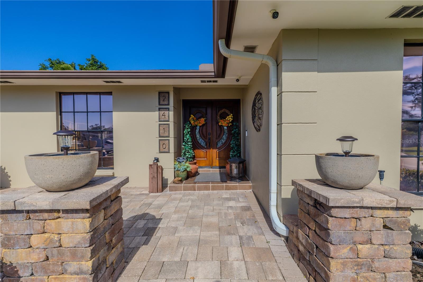 A welcoming front entry showcases a wide paver walkway framed by stone columns with accent lighting, leading to striking custom double wood doors imported from Malaysia beneath a covered entry for an elegant and secure arrival.