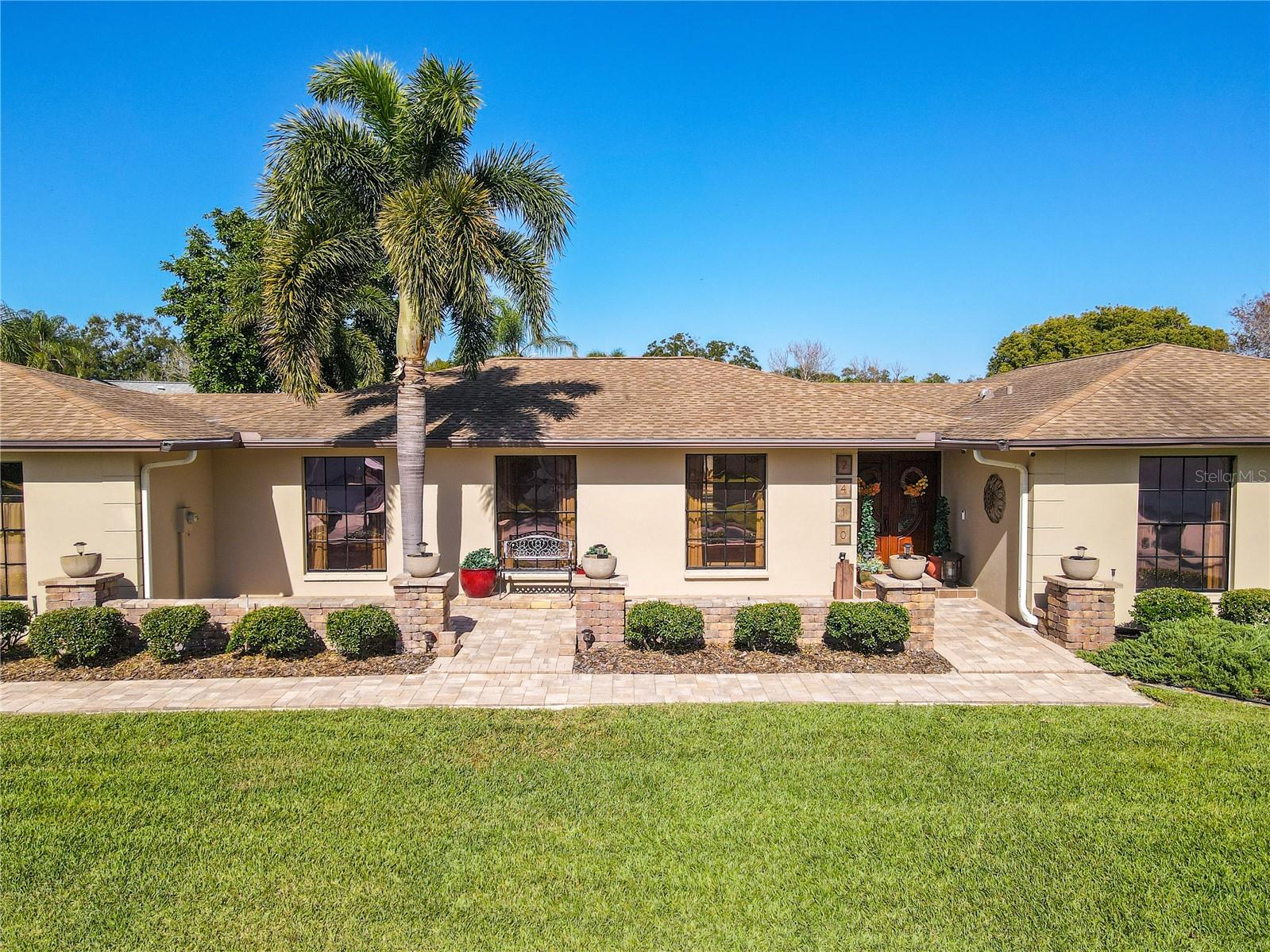 A wide, sunlit front elevation showcases manicured lawn, mature palm trees, stone-accented planters, and an inviting paver walkway leading to the welcoming entry.