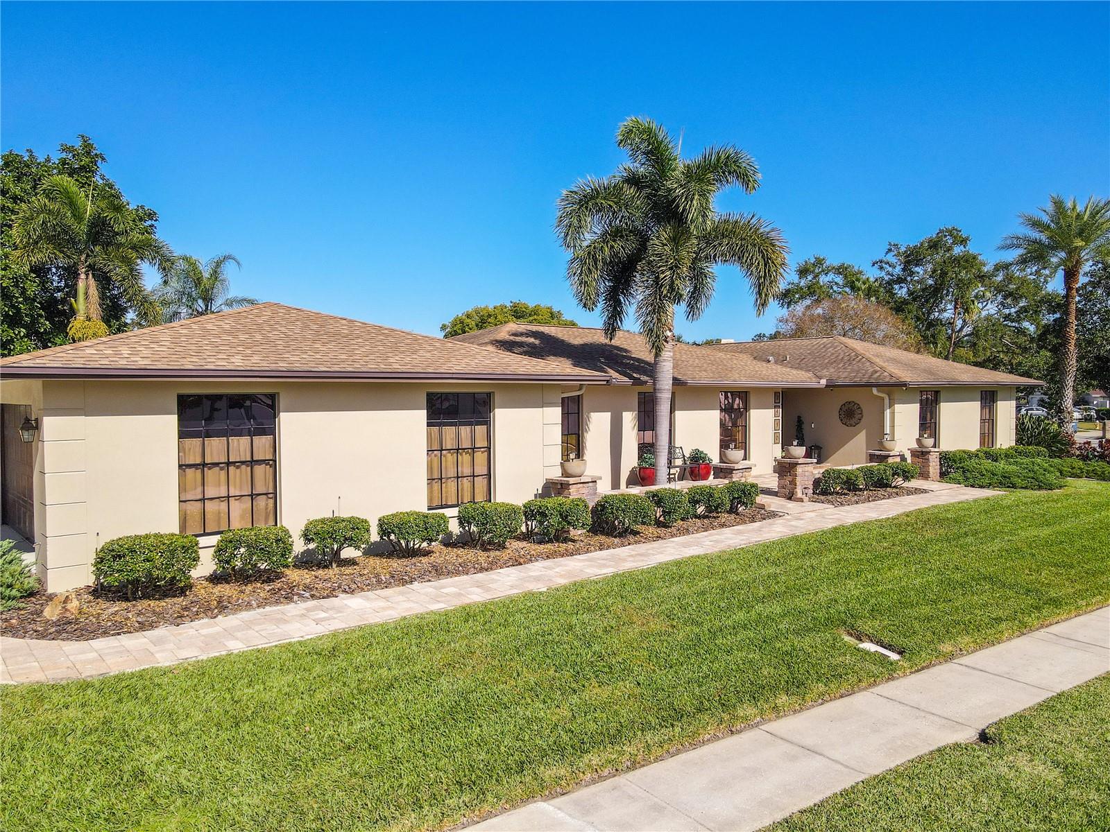 A sweeping street-side view highlights the home’s long, elegant façade with manicured hedges, mature palms, stone-accented planters, and a continuous paver walkway set against a lush green lawn and clear blue sky.