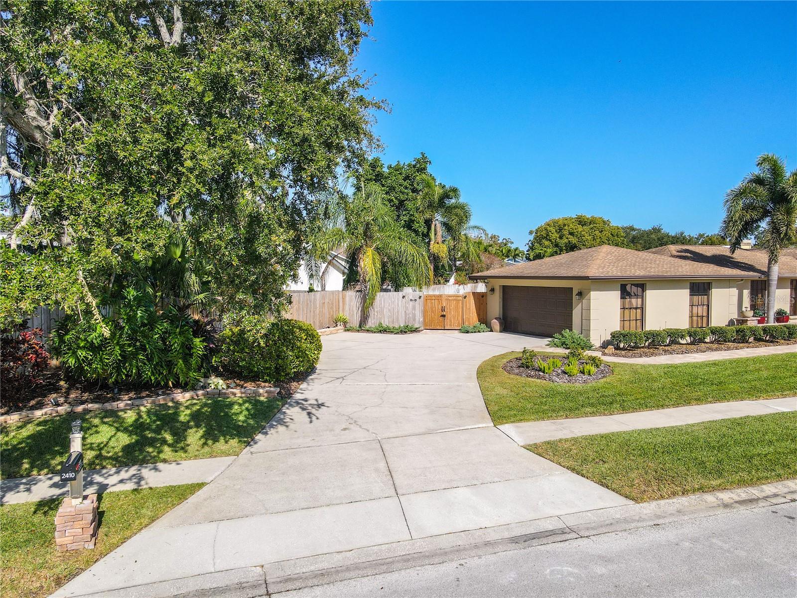 A wide-angle exterior view highlighting the home’s generous curved driveway, side-entry garage, double gate entry to the back yard, sidewalks, and neatly trimmed tropical landscaping that frames the property with excellent curb appeal and a clean, welcoming streetscape.