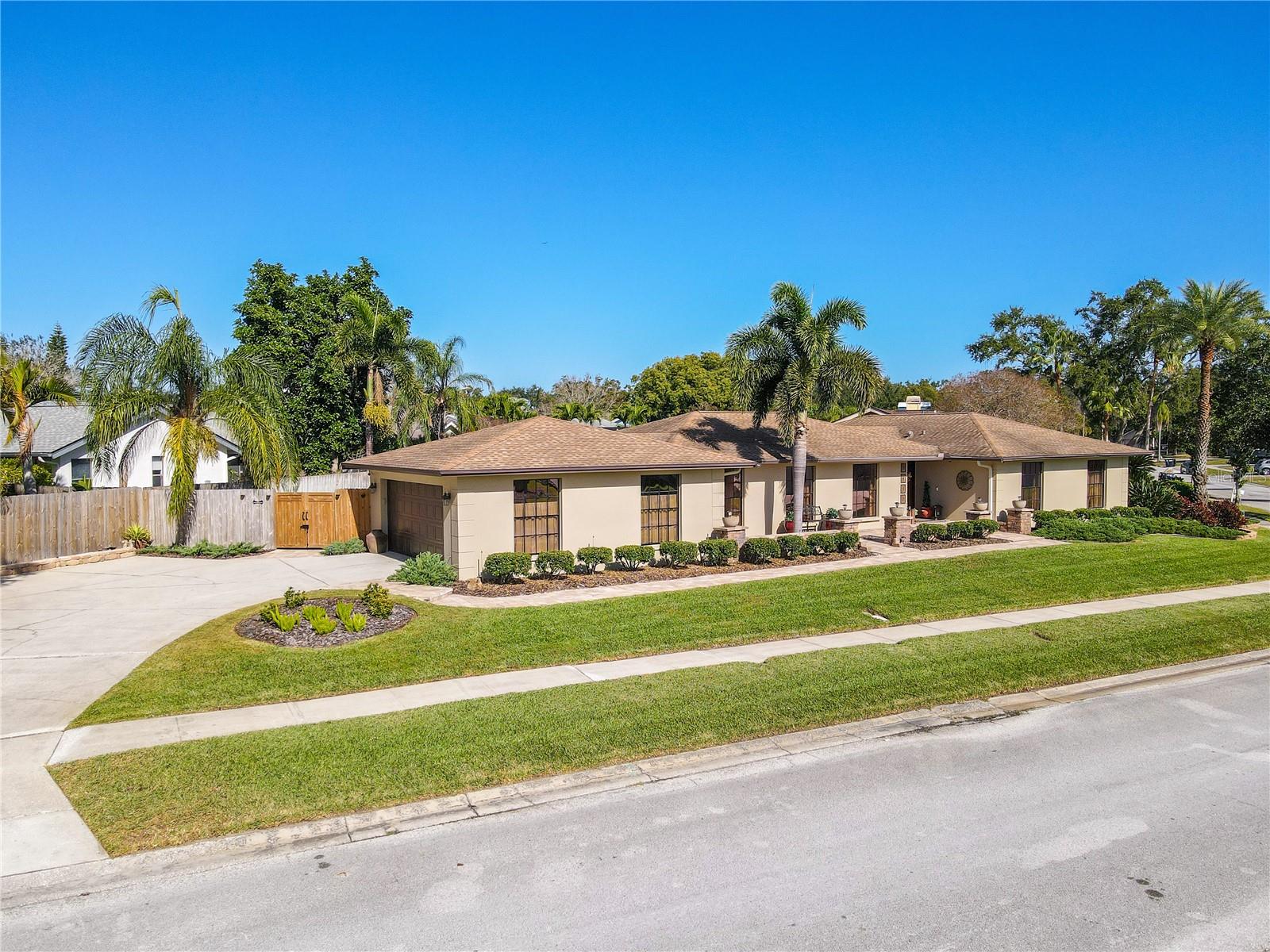 An expansive curbside aerial showcases the home’s wide frontage with a circular driveway, lush green lawn, mature palms, privacy fencing, and a long, paver-lined walkway leading to the welcoming front entry, all set within a quiet, established neighborhood.
