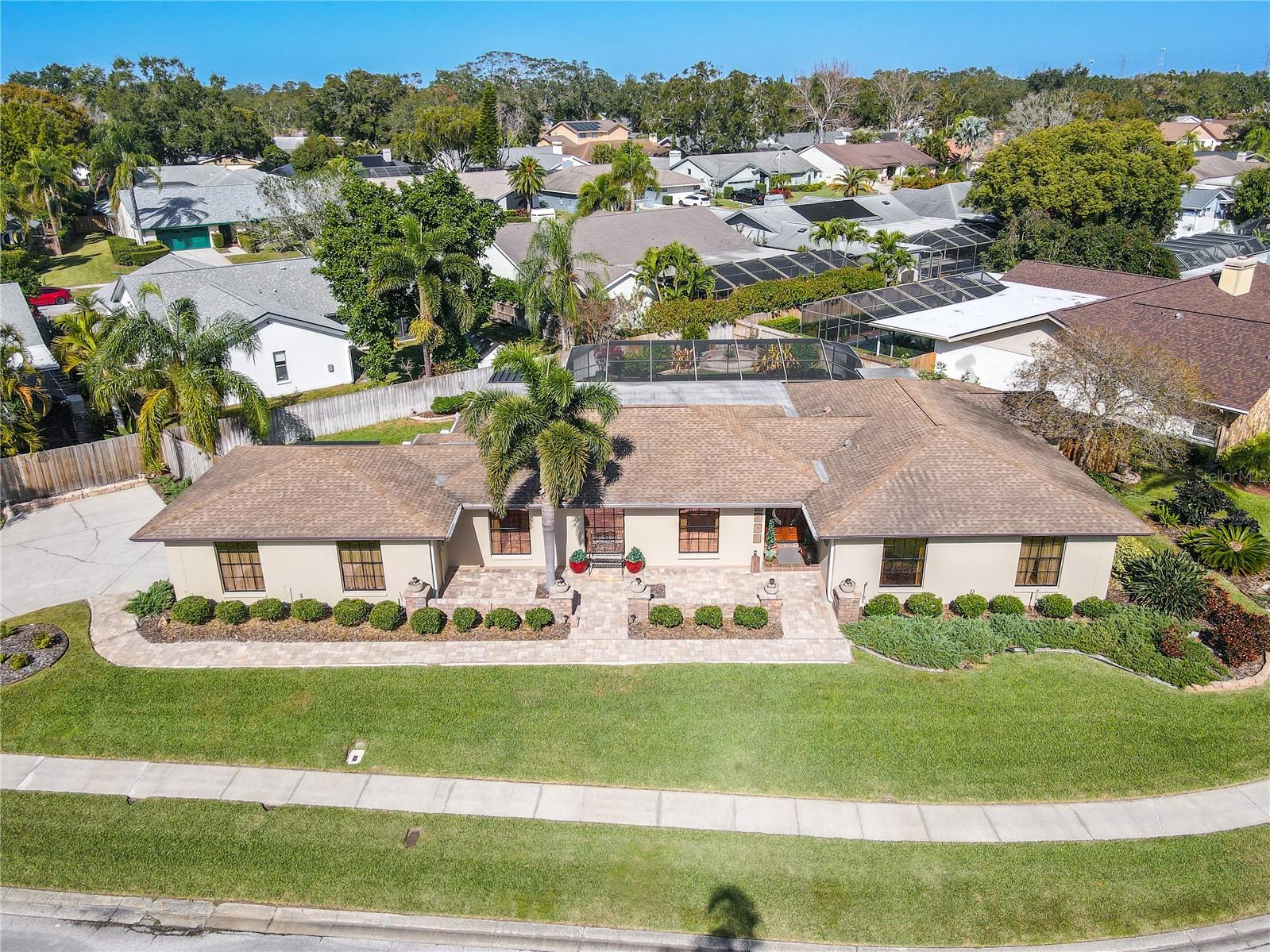 A straight-on aerial highlights the home’s broad, symmetrical façade with manicured hedges, mature palms, and an elegant paver walkway spanning the full frontage, set within a quiet neighborhood framed by lush tree canopy beyond.