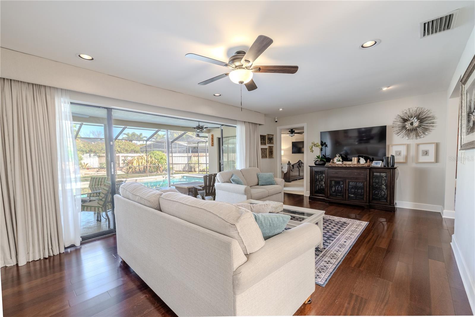 Spacious family room featuring rich hardwood floors, recessed lighting, and a ceiling fan, with expansive sliding glass doors that open to the screened lanai/pool area, creating a seamless indoor-outdoor living experience.