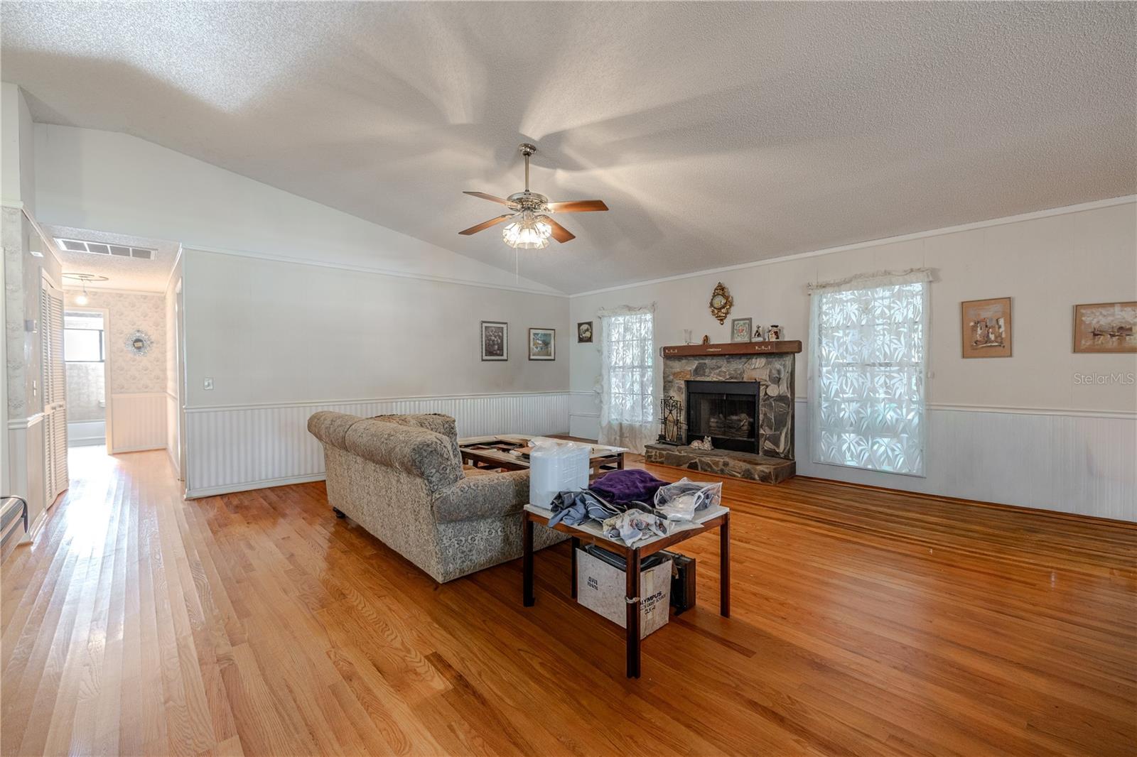 Expanded view of the open living space showcasing gleaming hardwood floors, wall papered wainscoting, ceiling fan lighting, and a cozy stone fireplace with plenty of room and entry to the bedrooms.