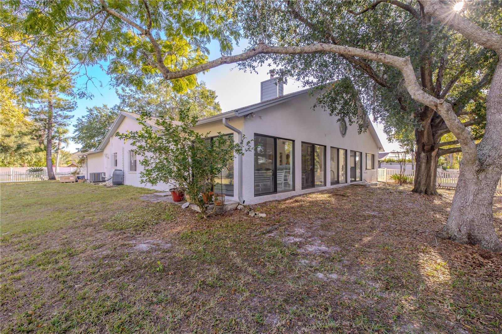 Spacious backyard view showcasing the home’s long wall of sliding glass doors along the enclosed Florida room, set beneath mature shade trees with a fenced yard and plenty of room to relax or entertain.