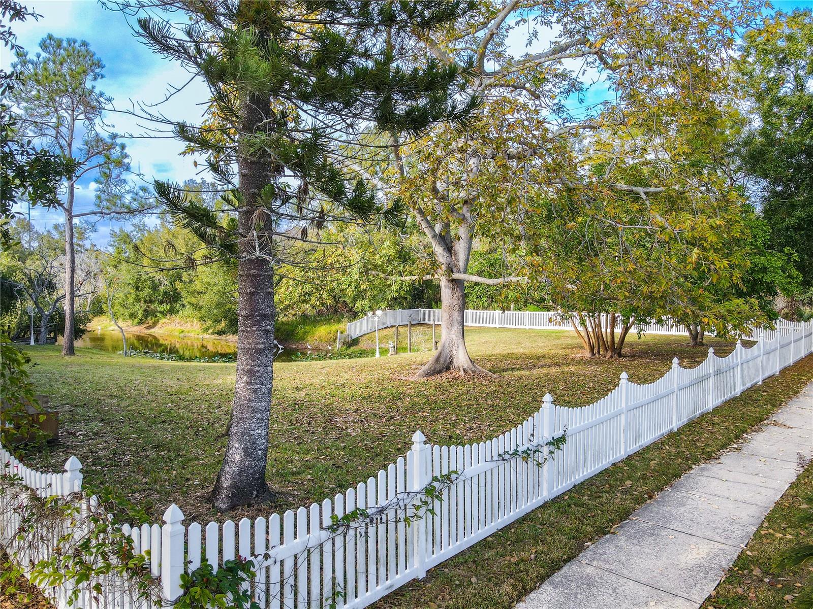 Front/side yard view featuring a charming white picket fence, expansive green space, and mature shade trees with glimpses of the peaceful private pond beyond.