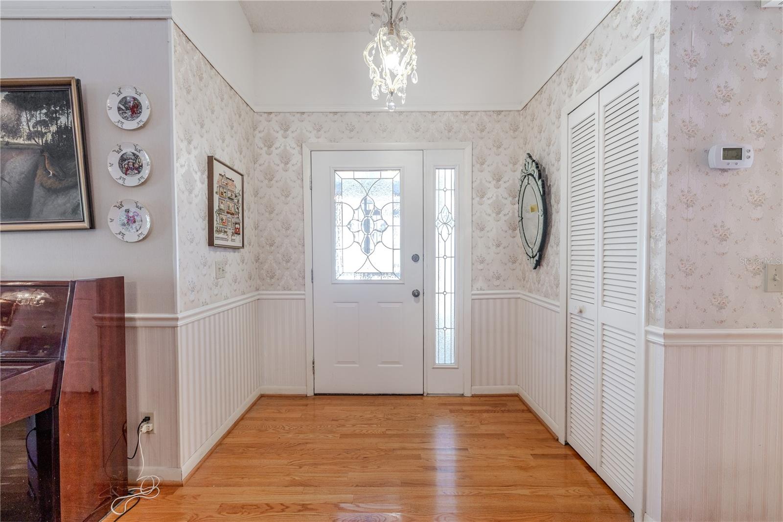 Bright entry hallway form a gated front patio with warm hardwood floors, white walls, a ceiling light fixture, and a front door with glass sidelights, plus nearby coat closet.