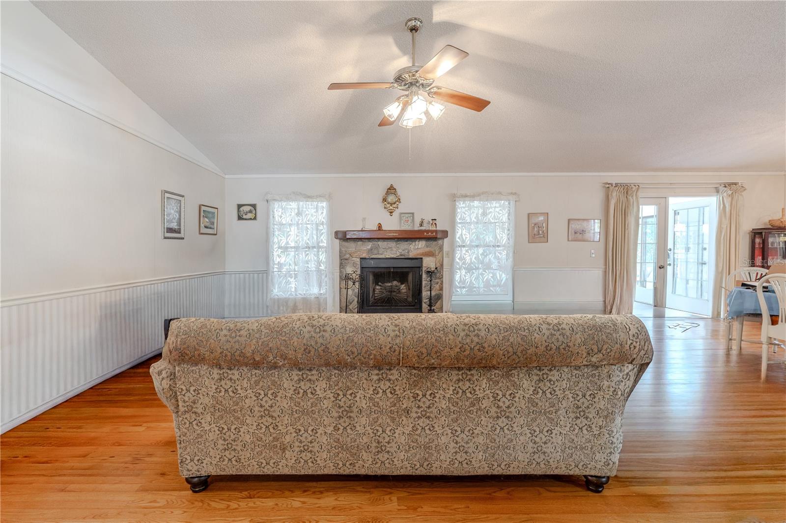 Spacious open vaulted ceiling 20.5 X 17.2 living area with ample natural light, wall papered wainscoting, gleaming hardwood floors, a ceiling fan, and a stone-accent fireplace centered between two bright windows, plus French doors leading to the large, open family room.