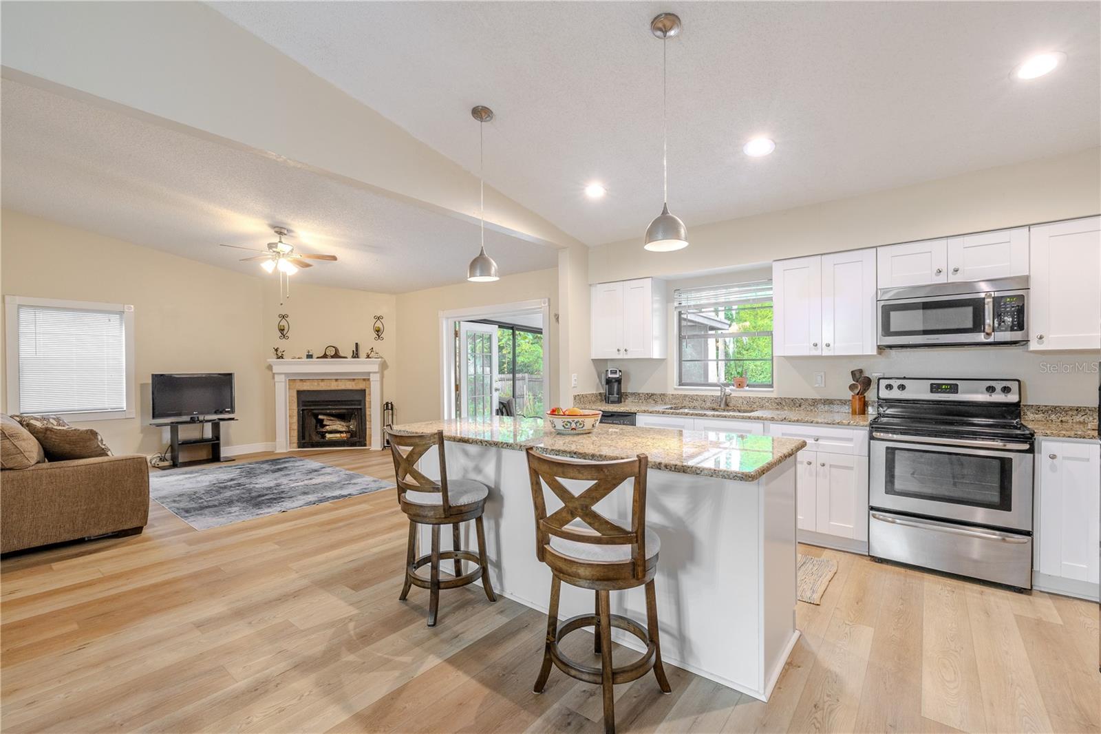 Bright open-concept living and kitchen space featuring a large granite island with seating, white cabinetry and stainless appliances, pendant and recessed lighting, wide Craftsman-style casing/trim on window and french doors with decorative corner rosette blocks and a cozy wood burning fireplace with french doors to the screened in porch.