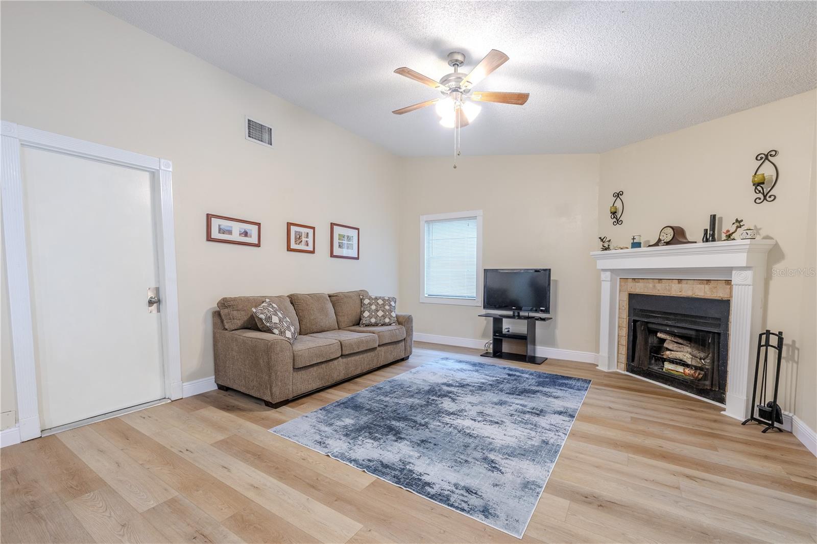 Comfortable living room with garage access, featuring wide-plank vinyl wood flooring, a ceiling fan, and a charming wood burning fireplace with mantel, creating a warm gathering space with plenty of room to relax.