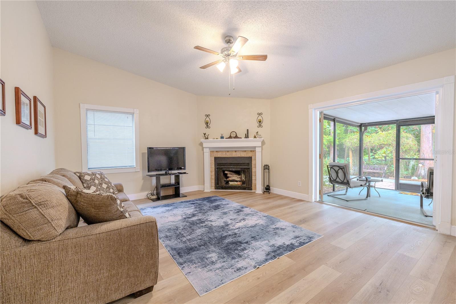 Bright living room with wide-plank vinyl wood flooring, vaulted ceiling and ceiling fan, centered around a cozy fireplace, with a large opening to the screened porch for seamless indoor-outdoor flow.