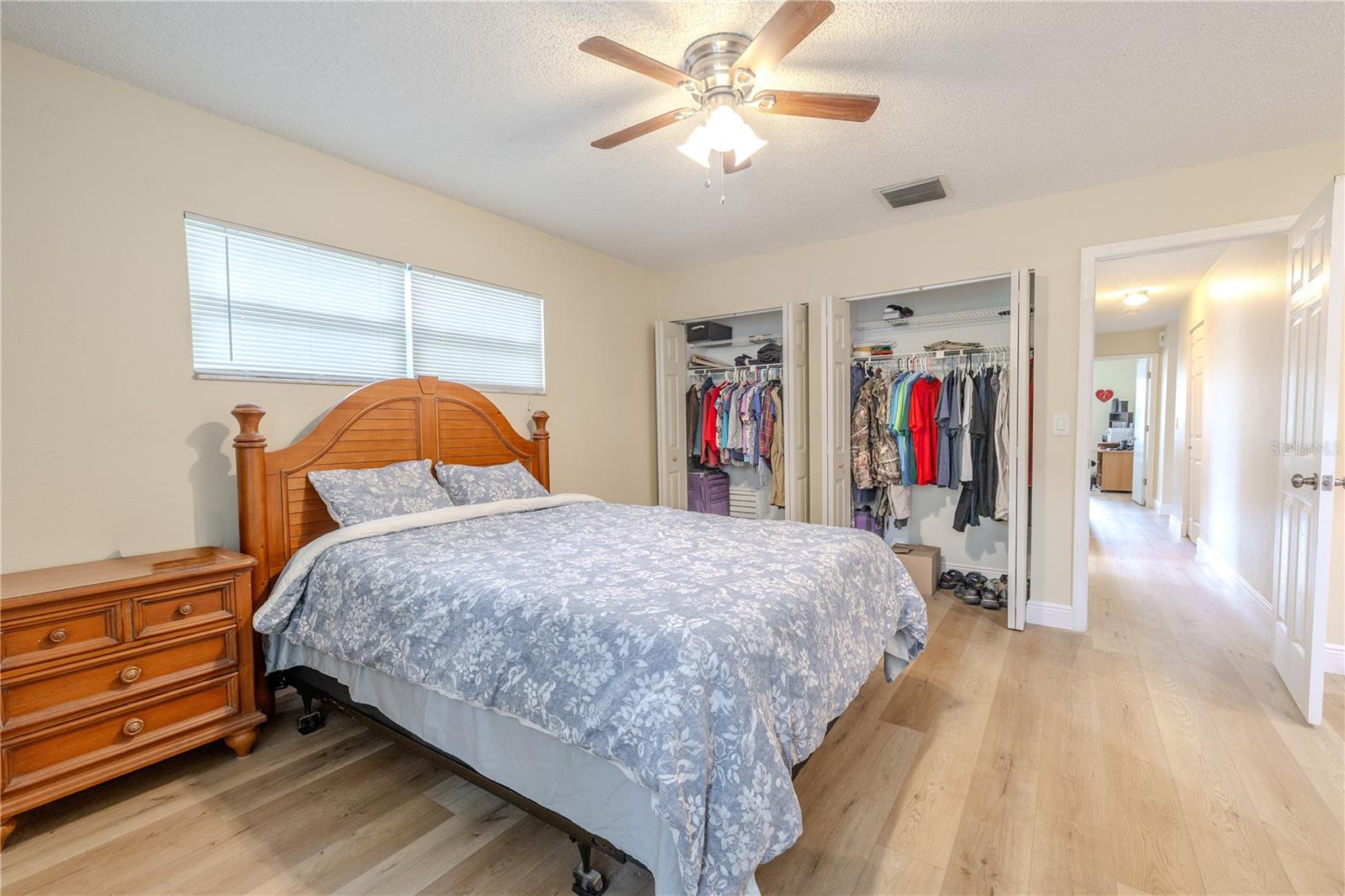 Additional view of the spacious 17 X 14 ft. primary bedroom featuring wide-plank vinyl wood flooring, 5" wood base boards, dual his and her built in closed, 6 panel door, a ceiling fan, and bright natural light from multiple windows for a calm, comfortable retreat.
