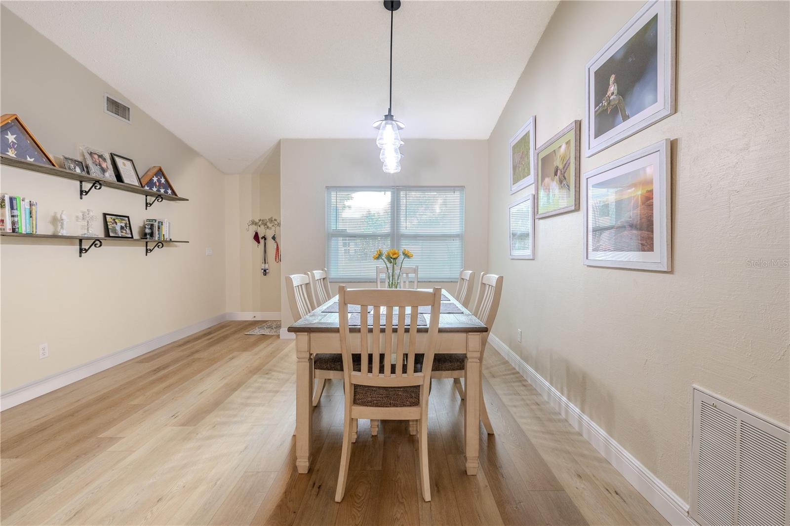 Full view of the spacious dining room with vaulted ceiling, wide-plank vinyl wood flooring, and a modern pendant light, anchored by a large window that brings in abundant natural light.