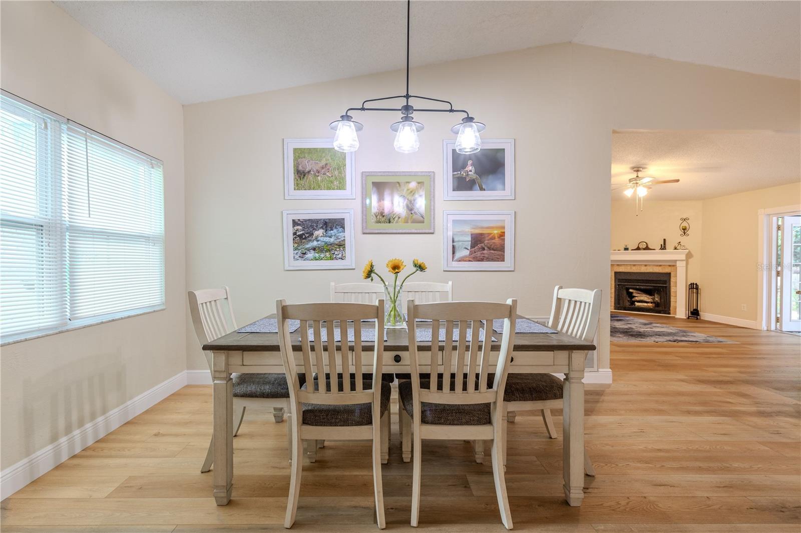Open dining space with vaulted ceiling, wide-plank vinyl wood flooring, and contemporary pendant lighting, seamlessly connecting to the living room with a cozy wood burning fireplace and french doors beyond.