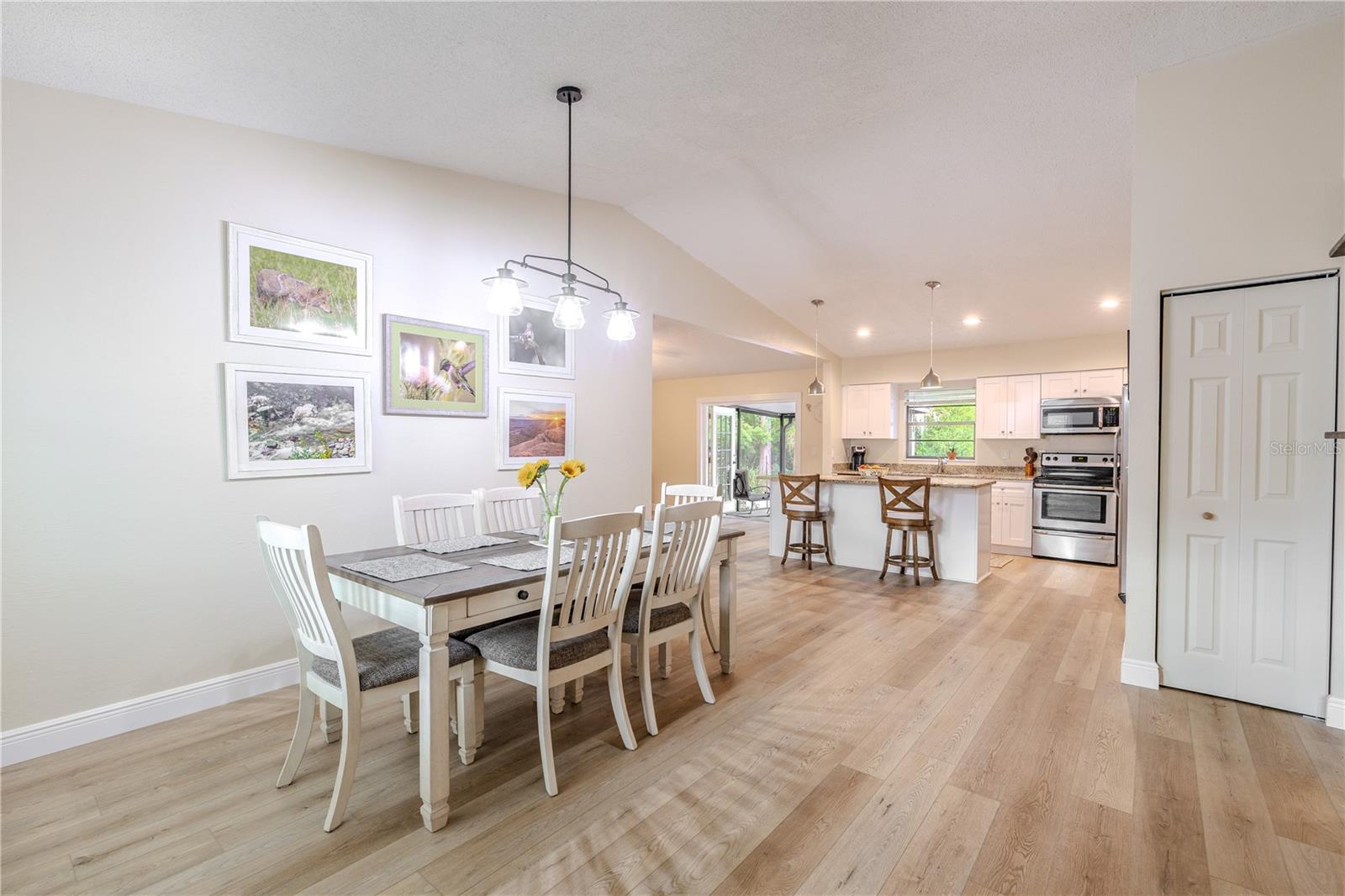 Spacious open layout showcasing the dining area flowing into the updated kitchen, featuring vaulted ceilings, wide-plank vinyl wood flooring, coat closet, pendant lighting, a breakfast bar, and stainless appliances.