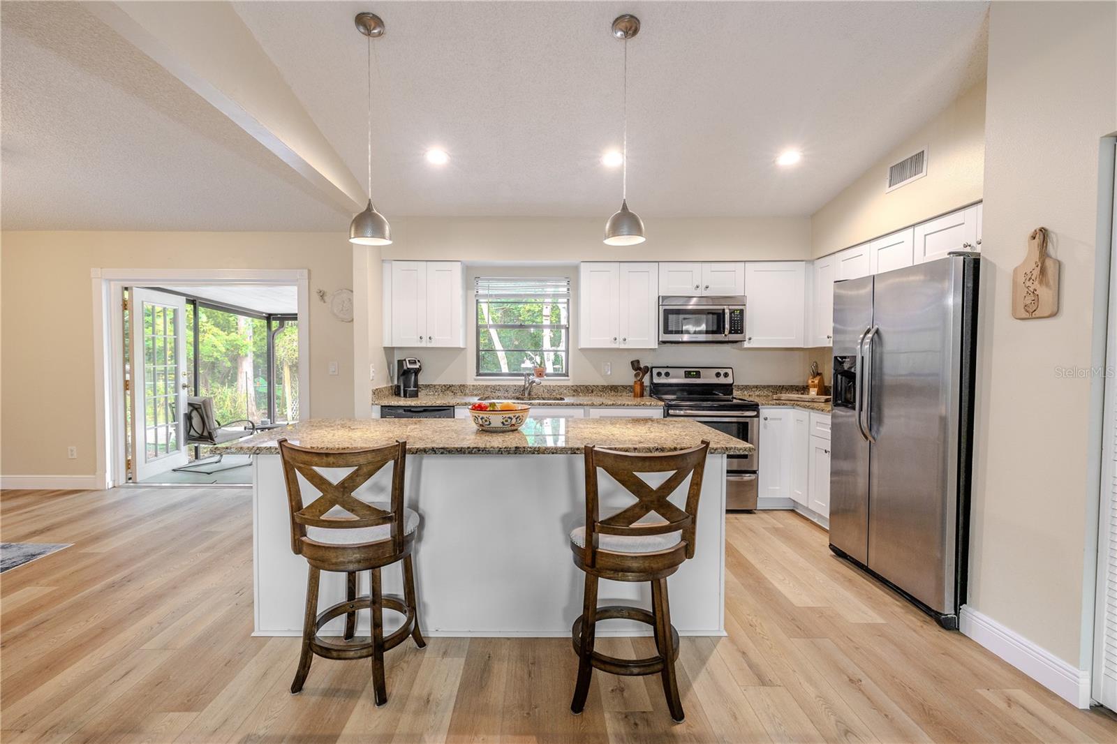 Bright, modern kitchen with white wood cabinetry, granite countertops, stainless appliances, and a large center island with breakfast-bar seating, all opening seamlessly to the living areas and screened in room beyond.
