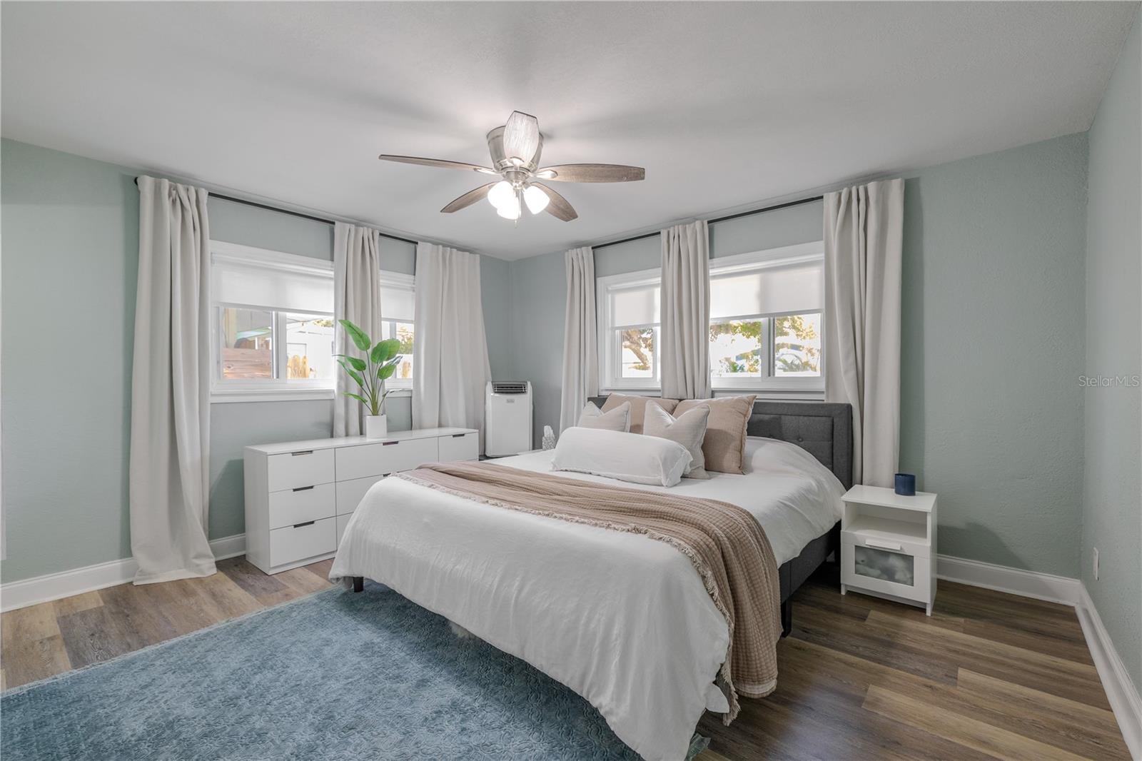 Serene primary bedroom features wide-plank vinyl flooring, a modern ceiling fan, and corner windows with soft drapery that fill the space with natural light.