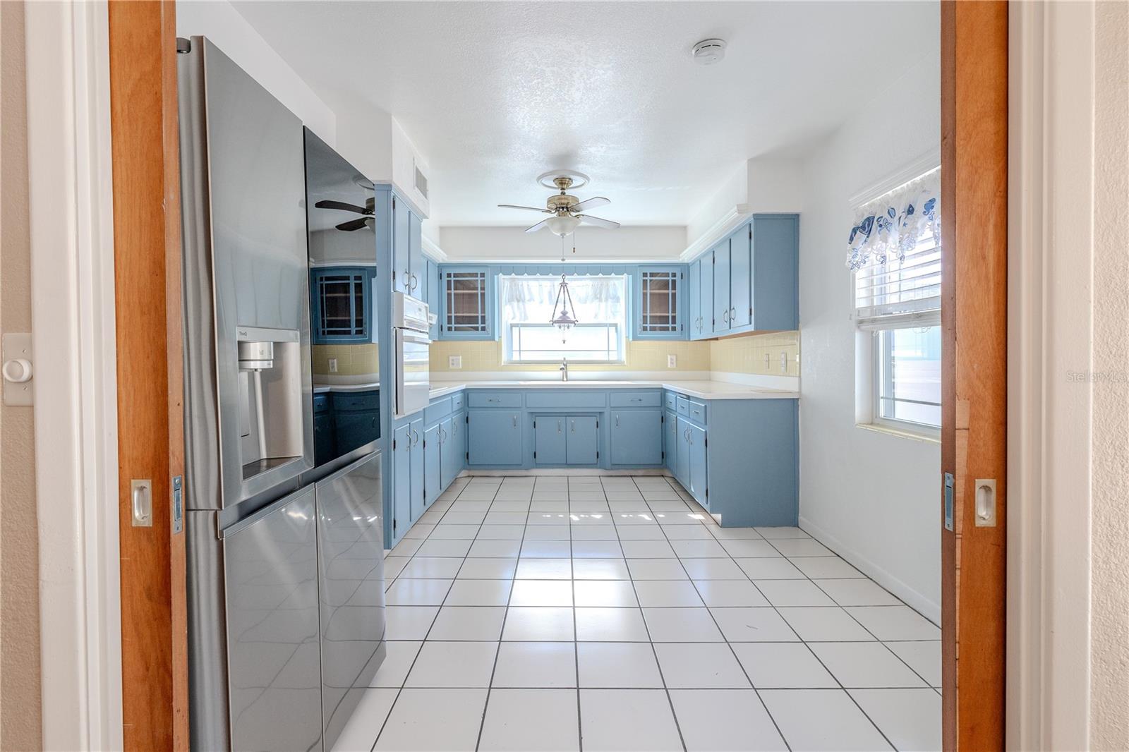 Bright, spacious eat in kitchen with charming solid wood blue cabinetry, abundant counter and storage space, tile flooring, and a stainless refrigerator, centered around a window over the sink for natural light.