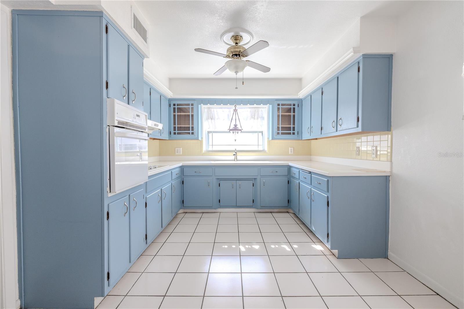 Direct view of the bright, spacious eat in kitchen with charming solid wood blue cabinetry, abundant counter and storage space, tile flooring, and a stainless refrigerator, centered around a window over the sink for natural light.